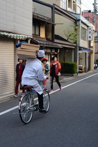 A Japanese man carrying two trays of food for delivery while riding a bicycle.