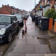 A Voi scooter parked on the pavement, narrowing the gap between a parked car and some bins