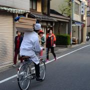 A Japanese man carrying two trays of food for delivery while riding a bicycle.